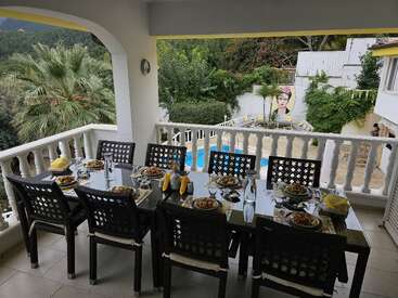Una acogedora mesa de comedor exterior para ocho personas con vistas a una piscina rodeada de exuberante vegetación, con un mural de Frida Kahlo al fondo. Ambiente relajante.