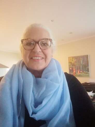 A smiling elderly woman with white hair and glasses is wearing a light blue scarf. She is indoors, with artwork and soft lighting in the background.