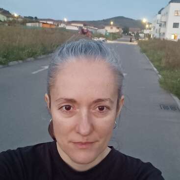 Eine Frau mit hellgrauem Haar macht ein Selfie auf einer verlassenen Straße in der Abenddämmerung. Im Hintergrund sind Häuser, Straßenlaternen und entfernte Hügel zu sehen.