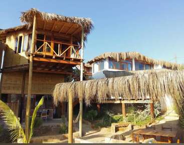 The image shows a rustic beachside building with thatched roofs, wooden supports, hammocks, outdoor seating, and tropical plants under a clear blue sky.