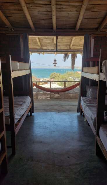 A cozy hostel room with bunk beds opens to a balcony. A hammock overlooks a stunning ocean view under a bamboo roof, inviting relaxation and tranquility.