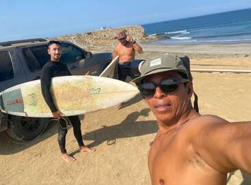 Three men pose for a selfie by the beach. One holds a surfboard, wearing wetsuit. A vehicle and ocean waves are visible in the sunny background.