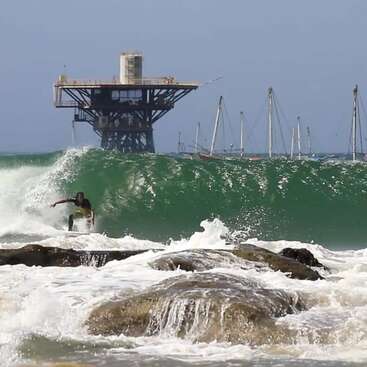A surfer rides a powerful green wave near rocky shore. In the background, an offshore oil platform and several sailboats are visible under a clear sky.