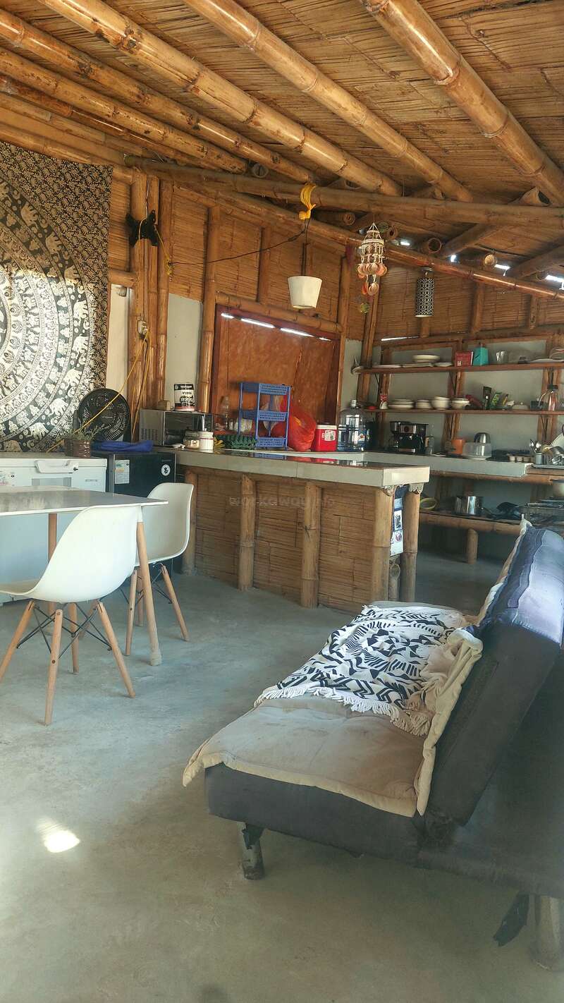 This cozy room features bamboo walls and ceiling, an eclectic kitchen, white chairs, a sofa with patterned blanket, open shelves, and a decorative tapestry.