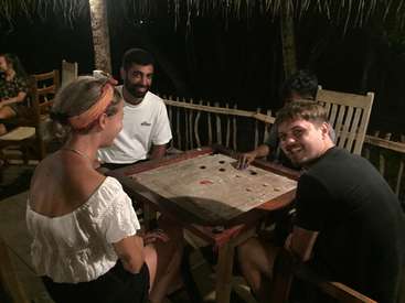 The image depicts four individuals seated around a wooden table, engaged in a game of carrom, with a serene outdoor setting at night.
