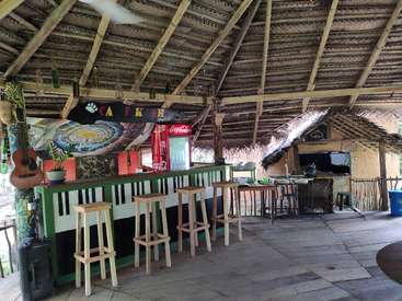 The image depicts a rustic bar with a thatched roof, featuring a counter with stools, a Coca-Cola fridge, and a TV, surrounded by a tropical atmosphere.