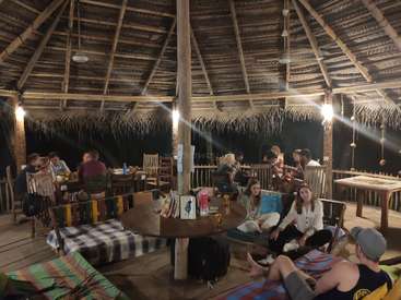 The image depicts a spacious, open-air dining area with a wooden floor and roof, featuring tables, chairs, and a group of people gathered in a relaxed atmosphere.