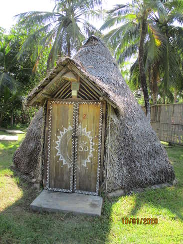 The image depicts a small, rustic hut with a thatched roof and wooden door, situated in a tropical setting surrounded by palm trees and lush greenery.