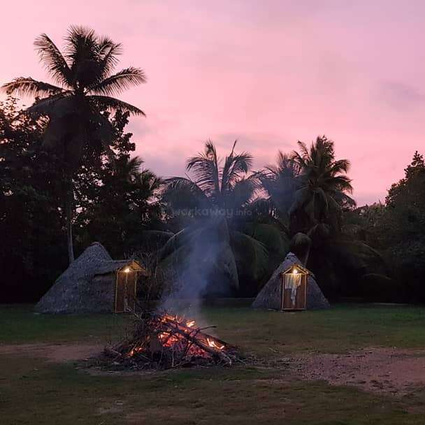 A serene sunset scene with two thatched huts, glowing lamps, tall palm trees, and a crackling bonfire in the center, surrounded by lush greenery.