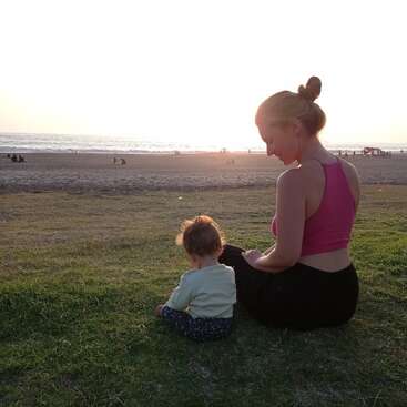 A imagem retrata uma cena serena de uma mulher e um bebê sentados na grama, olhando para a praia, com o sol se pondo ao fundo, criando uma atmosfera acolhedora.