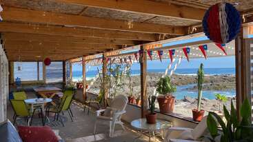 Un patio couvert confortable surplombe l'océan, décoré de guirlandes rouges, blanches et bleues. On y trouve des plantes en pot, des chaises, des tables et des décorations festives suspendues.