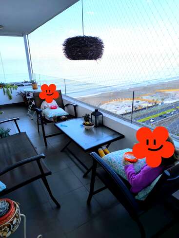 Deux personnes aux visages recouverts d'autocollants de fleurs sont assises sur un balcon avec vue sur l'océan et dégustent des collations. Des plantes, une lampe suspendue et un paysage côtier complètent la scène.