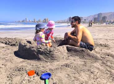 Un homme et deux enfants portant des chapeaux jouent dans le sable à la plage, construisant un château de sable avec des jouets, avec des bâtiments urbains et l'océan en arrière-plan.