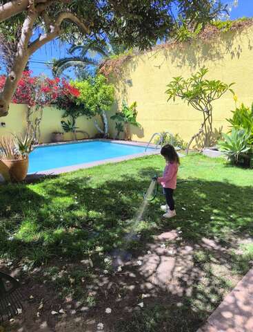 Une jeune fille arrose la pelouse d'un jardin ensoleillé, entouré de verdure luxuriante, de fleurs colorées et d'une piscine adossée à un mur jaune.