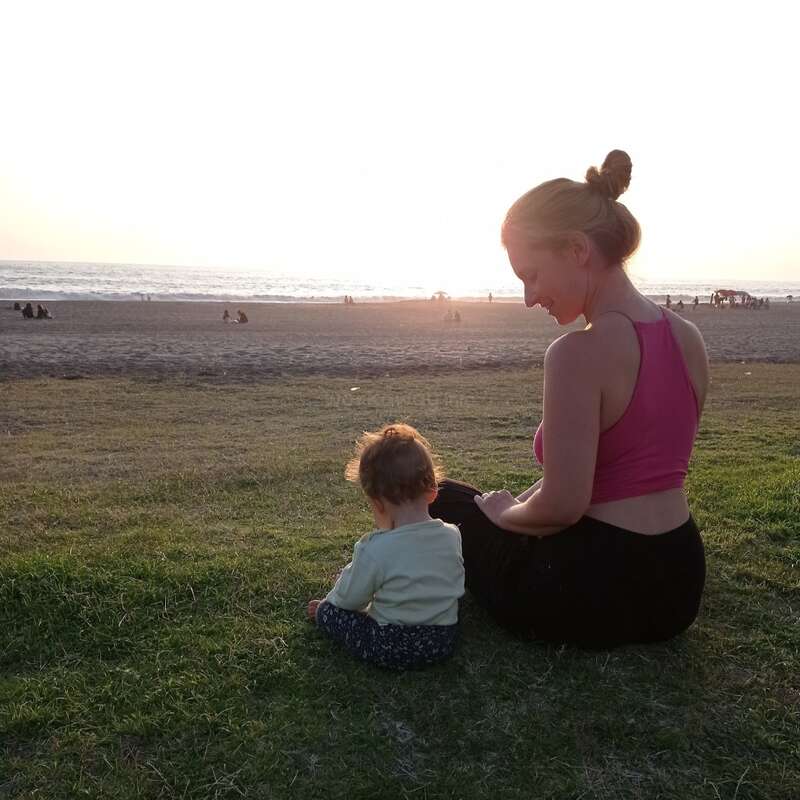 L'image représente une scène sereine d'une femme et d'un bébé assis sur l'herbe, regardant la plage, avec le soleil se couchant en arrière-plan, créant une atmosphère chaleureuse.