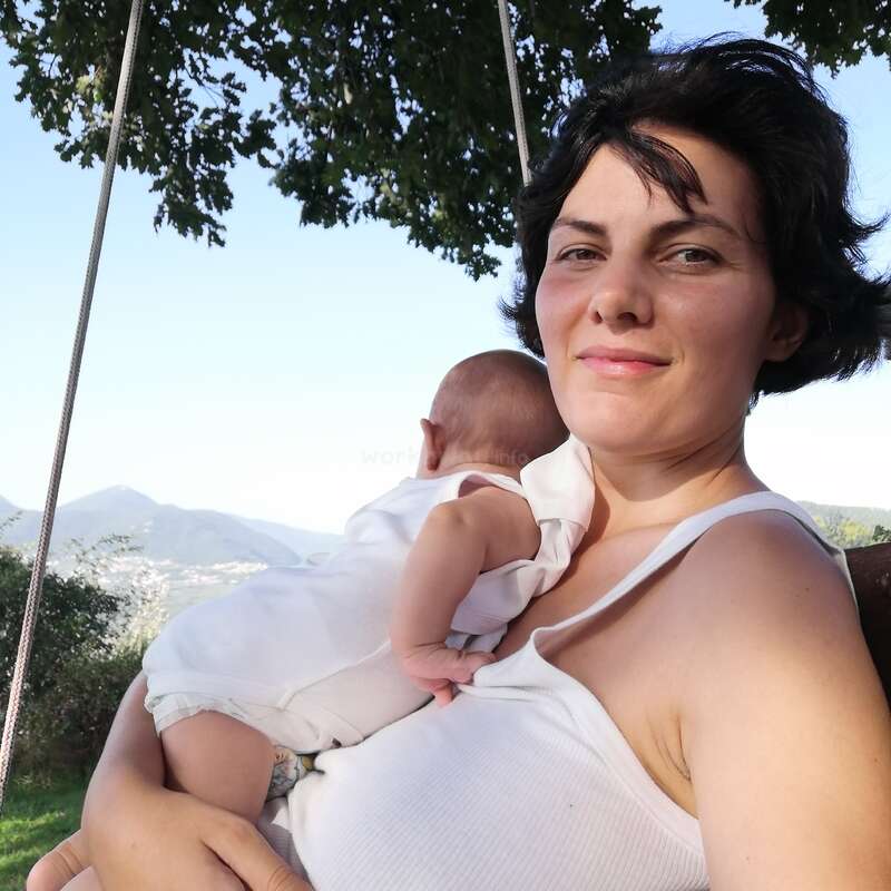 Das Bild zeigt eine Frau mit kurzen dunklen Haaren, die ein Baby auf ihrer Schulter trägt, vor einer ruhigen Landschaft mit Bäumen, Bergen und einem klaren blauen Himmel.
