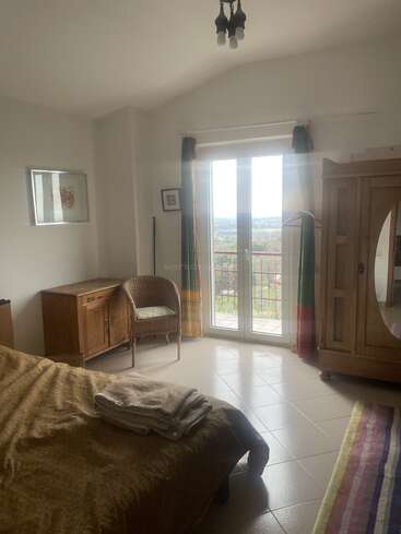 This cozy, sunlit bedroom features a bed with blankets, wicker chair, wooden wardrobe, desk, and French doors leading to a balcony with a scenic view.