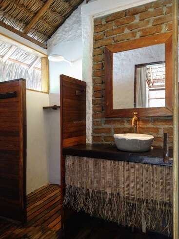 La imagen muestra un cuarto de baño rústico con lavabo, espejo y pared de ladrillo, en el que destacan una encimera de madera y una alfombrilla tejida, que desprenden un ambiente natural y terrenal.