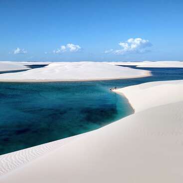 L'image représente un paysage désertique serein avec des dunes de sable blanc et une paisible étendue d'eau bleue, sur fond de ciel bleu clair.