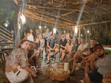 Un grupo de amigos se sientan juntos bajo un rústico tejado de paja por la noche, sonriendo, charlando y disfrutando de las bebidas en un ambiente cálido y festivo al aire libre.
