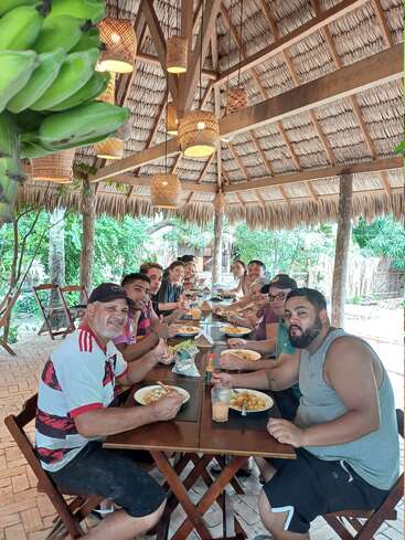 Un grupo numeroso de personas disfruta de una comida bajo un pabellón con tejado de paja, rodeado de naturaleza y decoración de madera, compartiendo risas y comida en un entorno tropical.