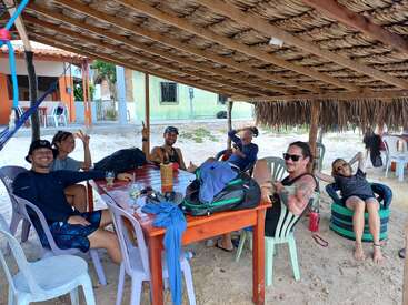 Seis personas se relajan bajo una cabaña con techo de paja en la playa, sentadas en sillas y neumáticos reciclados, sonriendo y descansando alrededor de una mesa con bebidas y mochilas.