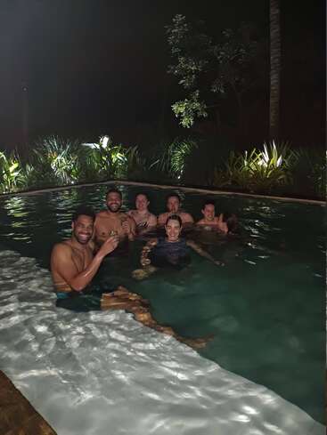 Un grupo de amigos disfruta de un baño nocturno en una piscina, sonrientes y relajados. La piscina está rodeada de vegetación, suavemente iluminada por luces.