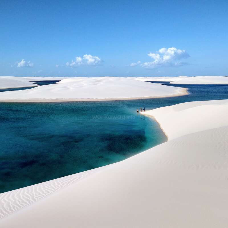 La imagen representa un sereno paisaje desértico con dunas de arena blanca y una tranquila masa de agua azul, sobre un cielo azul despejado.