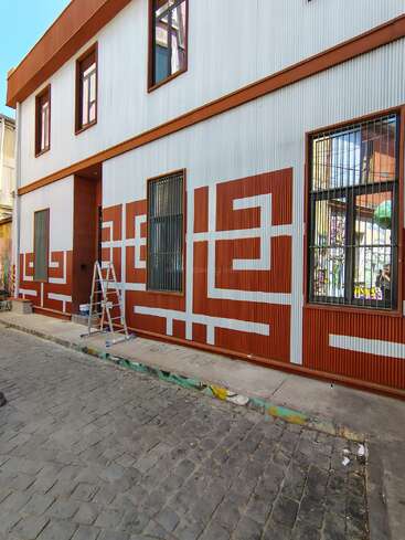 A building with a geometric mural in rust and white, barred windows, a ladder, paint supplies, cobblestone street, clear sky, and urban atmosphere.