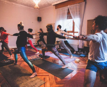 Das Bild zeigt eine Gruppe von Menschen, die in einem Raum mit weißen Wänden, Holzböden und einem großen Fenster auf Yogamatten stehend Yoga üben.