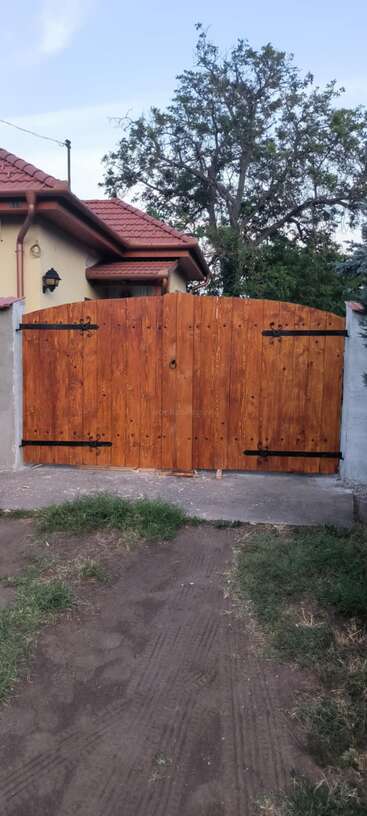 A freshly installed wooden gate stands between concrete walls, in front of a house with a red-tiled roof and a large leafy tree behind it.