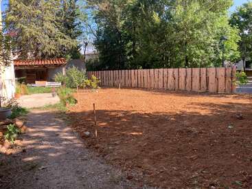 Una valla de madera delimita una parcela recién labrada. Árboles y vegetación rodean la zona, con una pequeña estructura y plantas a la izquierda.