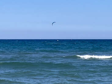 Une scène d'océan calme avec de douces vagues qui roulent vers le rivage. Le ciel est d'un bleu limpide et la voile d'un cerf-volant solitaire est visible au loin.