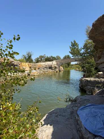 Ein ruhiger Fluss fließt unter einer Steinbrücke, umgeben von felsigen Klippen und Grün. Eine Person schwimmt, während eine blaue Decke auf den Felsen liegt.