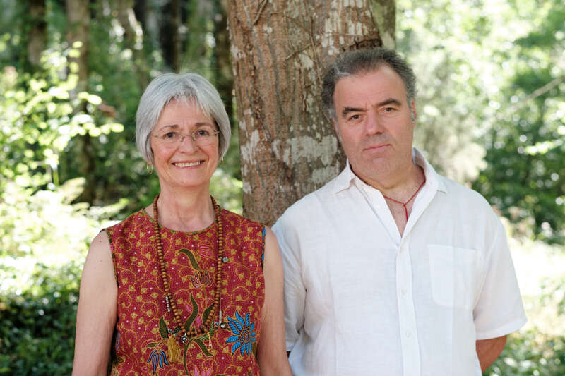 Eine ältere Frau und ein Mann stehen im Freien vor einem Baum. Die Frau lächelt, trägt eine Brille und ein gemustertes Kleid. Es ist ein sonniger Tag.