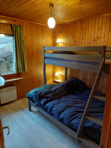 Esta acogedora habitación de madera cuenta con una litera gris con ropa de cama oscura, iluminación cálida, una ventana con cortinas verdes y suelos de madera clara, lo que crea un ambiente rústico.