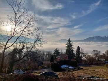 Une scène pittoresque avec une maison au toit rouge, des arbres dénudés, des pins, un brouillard brumeux, des montagnes enneigées et un soleil rayonnant derrière des nuages épars dans le ciel bleu.