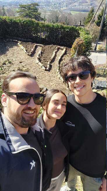 Three friends smile for a selfie outdoors with sunglasses, standing in a sunny garden. Behind them, raised garden beds form a heart shape. Trees and hills in background.