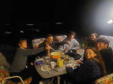 Seis amigos están reunidos alrededor de una mesa por la noche, disfrutando juntos de una fondue al aire libre. Sonríen, se ríen y comparten una cena acogedora y alegre.