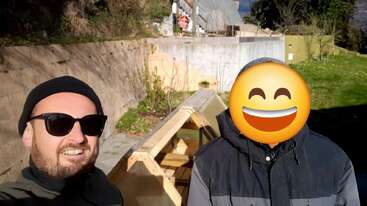 Dos personas están al aire libre junto a una estructura de madera; una sonríe a la cámara con gafas de sol, mientras que el rostro de la otra está cubierto por un emoji sonriente. Es un día soleado.
