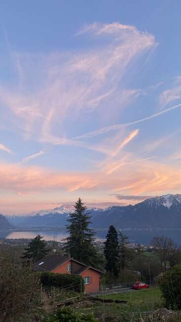 Un sereno cielo al atardecer con nubes en tonos pastel, montañas nevadas, un lago tranquilo, árboles de hoja perenne, una casa de tejado rojo y un coche rojo crean una apacible escena campestre.