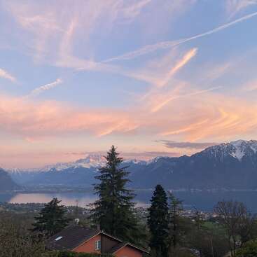 Un ciel serein au coucher du soleil, parsemé de nuages aux teintes pastel, des montagnes enneigées, un lac paisible, des arbres à feuillage persistant, une maison au toit rouge et une voiture rouge composent une scène champêtre apaisante.