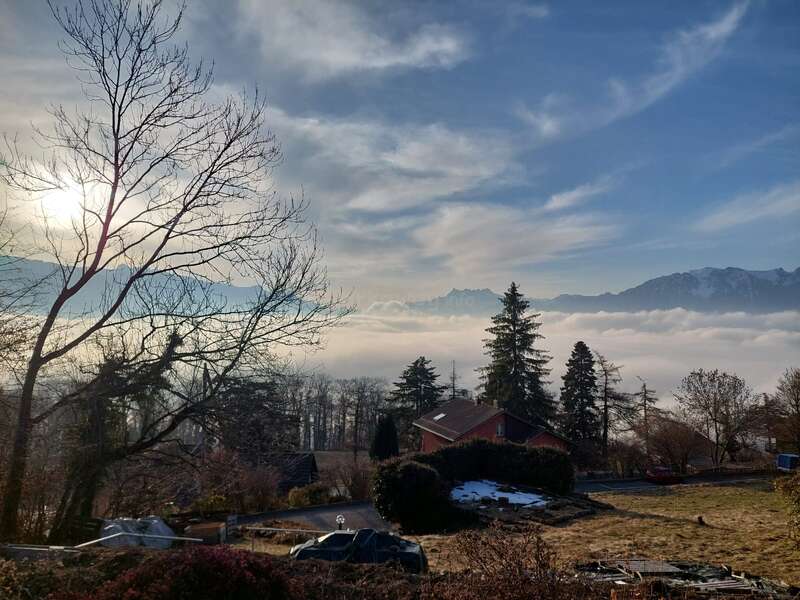 Una escena pintoresca con una casa de techo rojo, árboles desnudos, pinos, niebla brumosa, montañas nevadas y un sol resplandeciente detrás de nubes dispersas en el cielo azul.