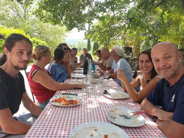 A imagem mostra um grupo de pessoas sentadas em uma mesa comprida, desfrutando de uma refeição em um ambiente externo sereno, cercado por árvores e vegetação exuberante.