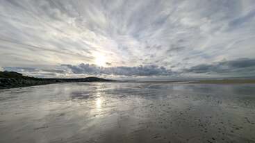 A wide, serene beach at low tide reflects dramatic, cloudy skies. The sun is partially hidden, casting soft light. Distant hills and calm waves enhance tranquility.