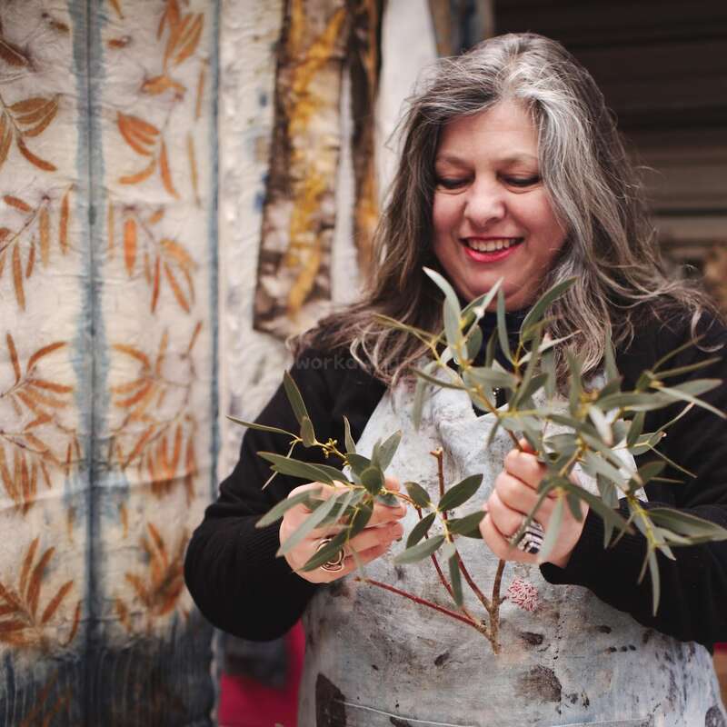 A smiling woman with gray hair holds leafy branches, wearing a paint-stained apron. Behind her hang textiles with natural leaf prints in earthy tones.