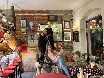 Three people are in a cozy, art-filled room with brick walls. Two stand near the wall; one relaxes on a couch, smiling and holding a phone.