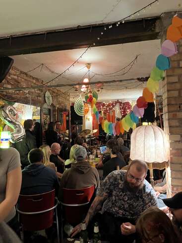 A cozy indoor gathering with people seated, listening to live music. Colorful decorations, string lights, and a vintage lamp create a festive, intimate atmosphere. Warm, social vibe.