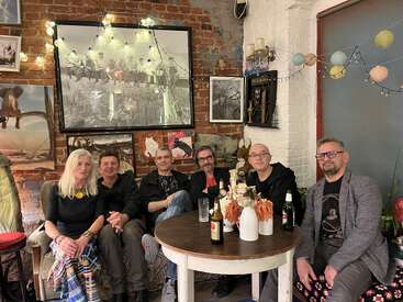 Six people sit together in a cozy, eclectic room with brick walls, artwork, string lights, drinks on the table, and a relaxed, friendly atmosphere.