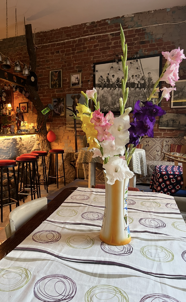 A rustic, cozy café features exposed brick walls, eclectic decor, a table centerpiece with colorful gladiolus flowers in a vase, high stools, and warm ambient lighting.